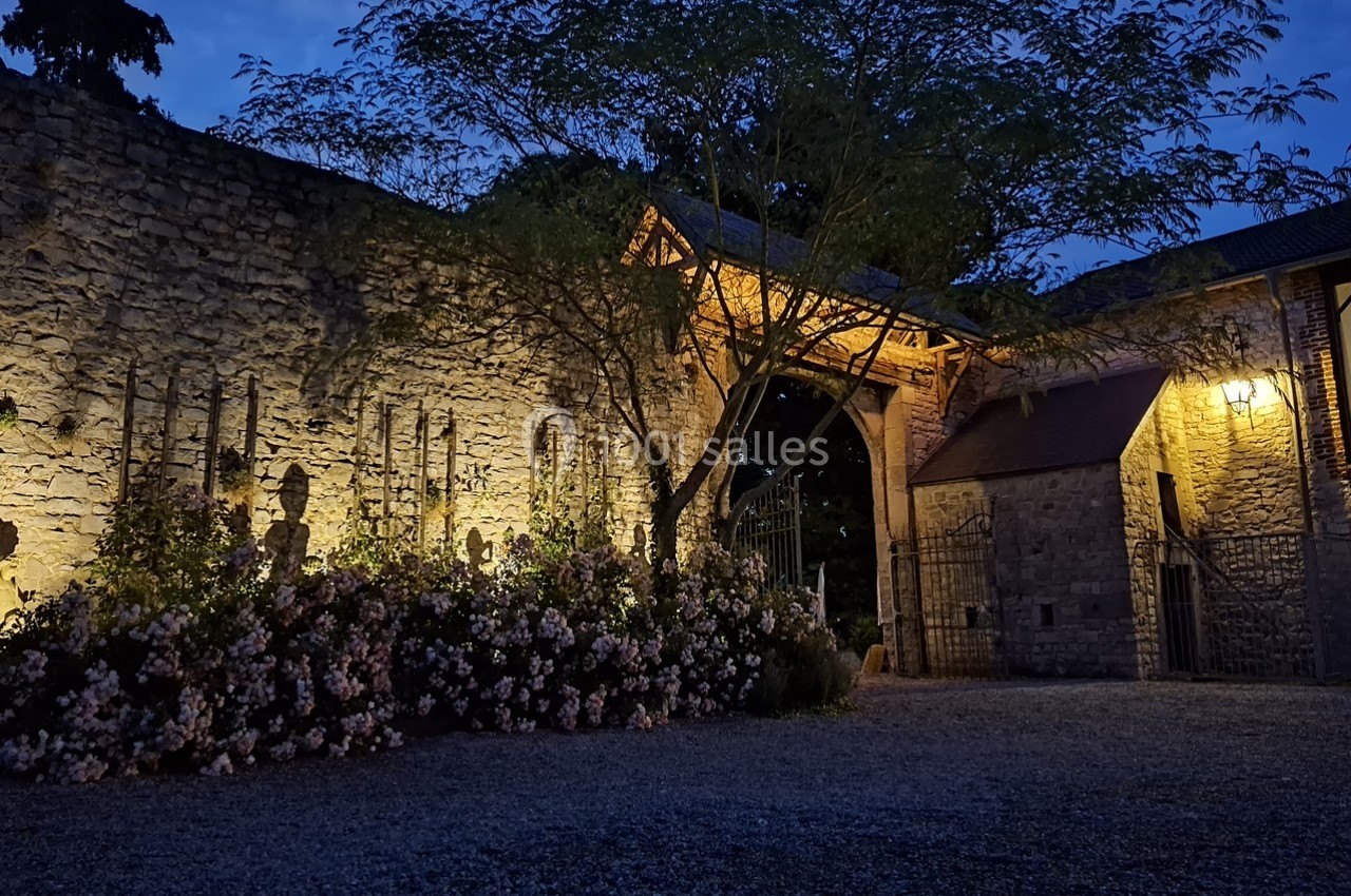 Entrée en pierre éclairée de nuit, entourée de fleurs et d'arbres, avec un portail en fer forgé.