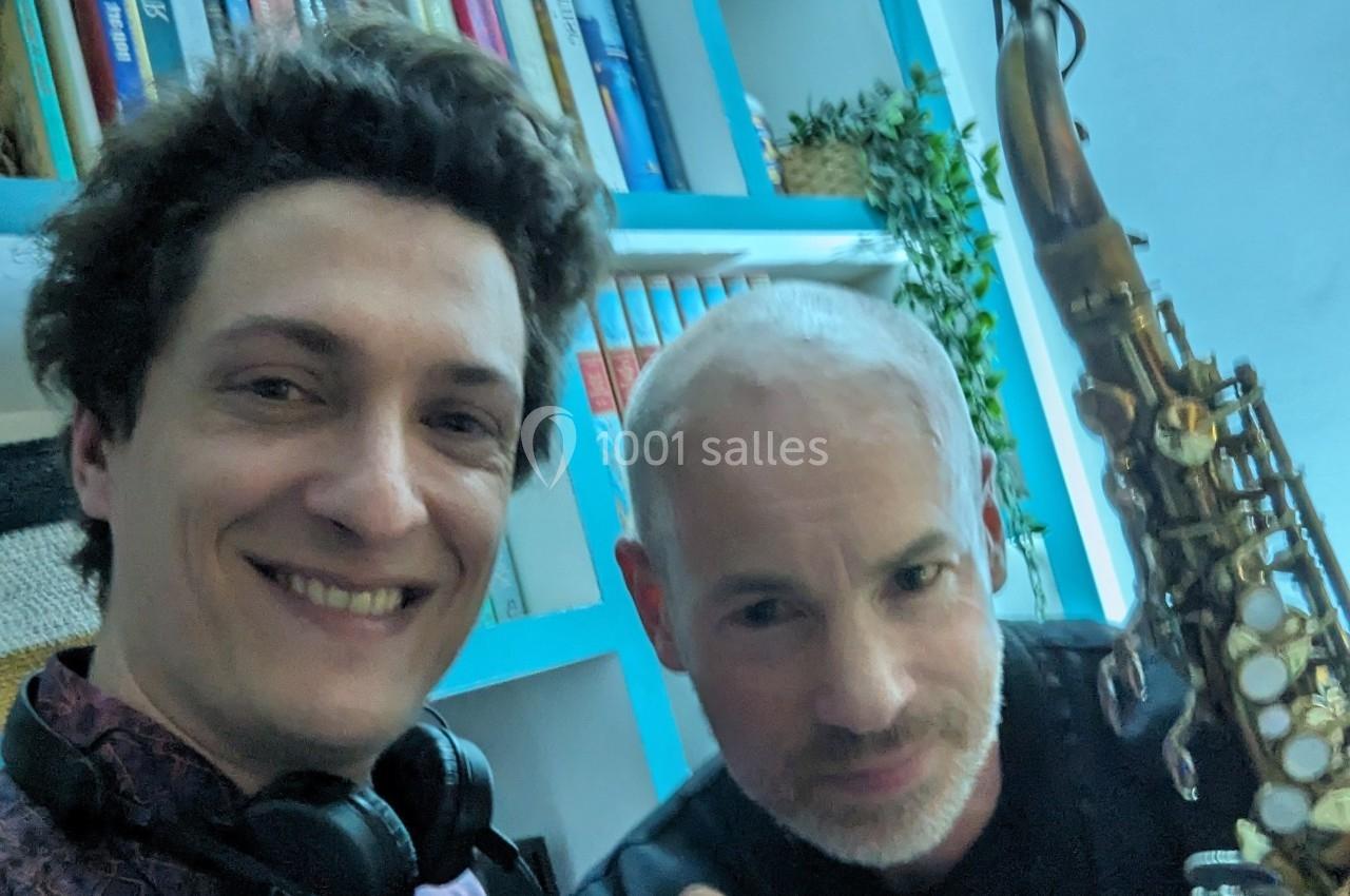 Deux hommes souriants posent devant une étagère bleue, l'un tenant un saxophone.