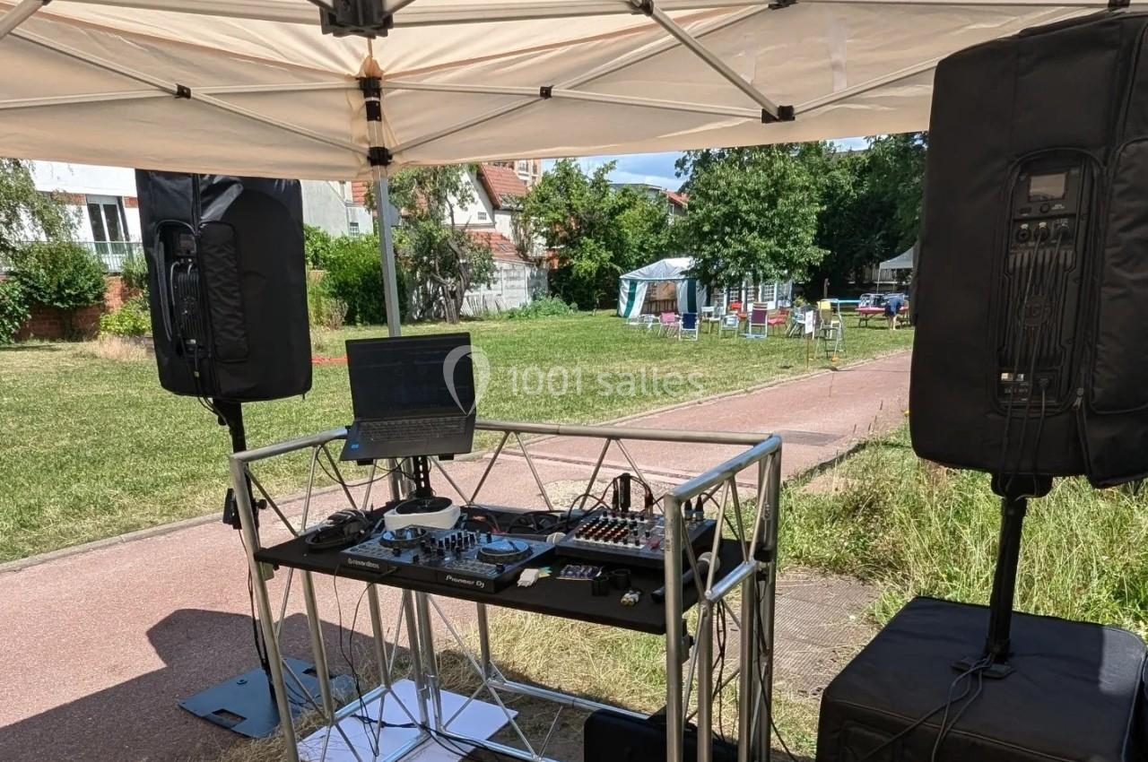 Équipement de sonorisation installé sous un abri, avec table de mixage, ordinateur et enceintes, dans un espace extérieur.