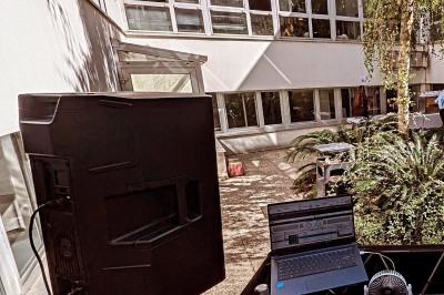 Un homme s'exprime au micro devant un public rassemblé dans une cour extérieure entourée de végétation.