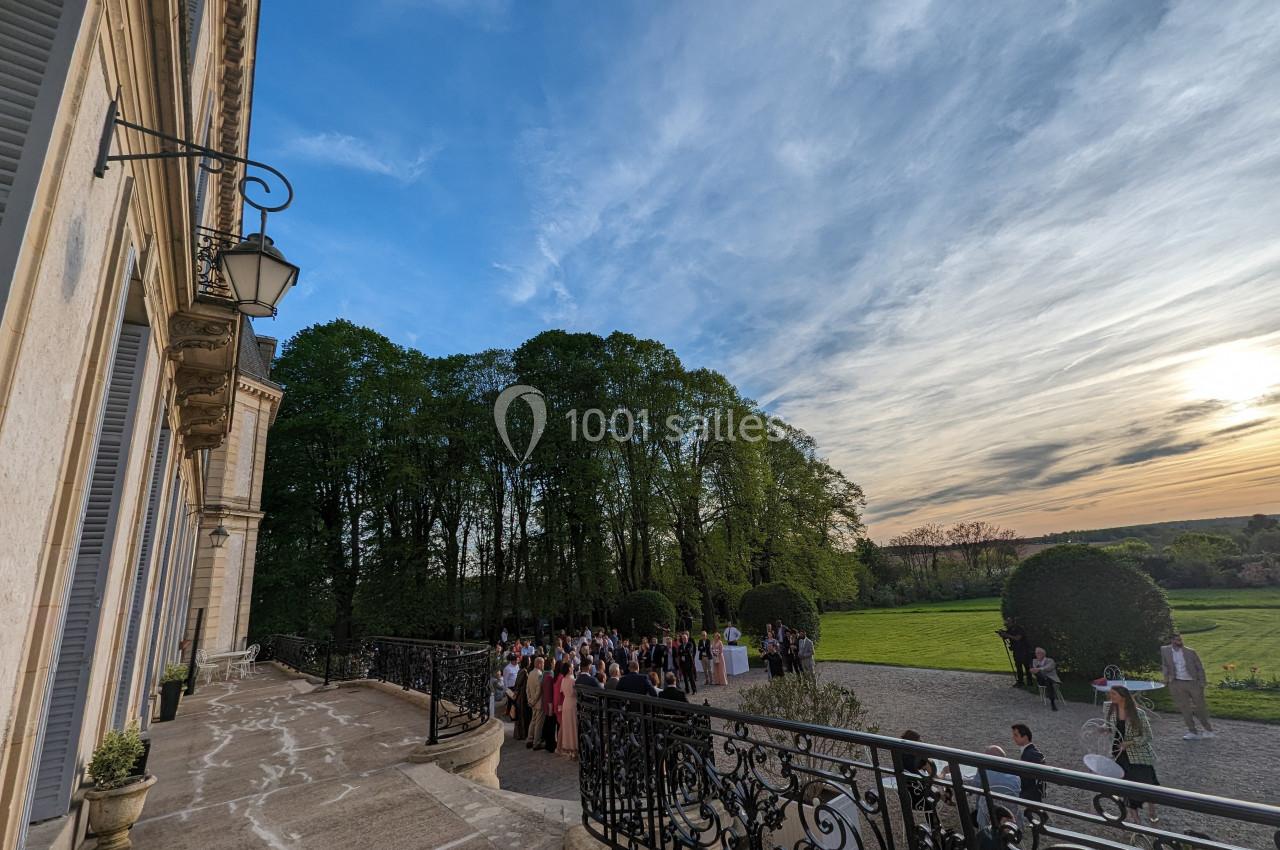 Groupe de personnes rassemblées sur une terrasse devant un bâtiment historique, avec un parc et un ciel au coucher du soleil.