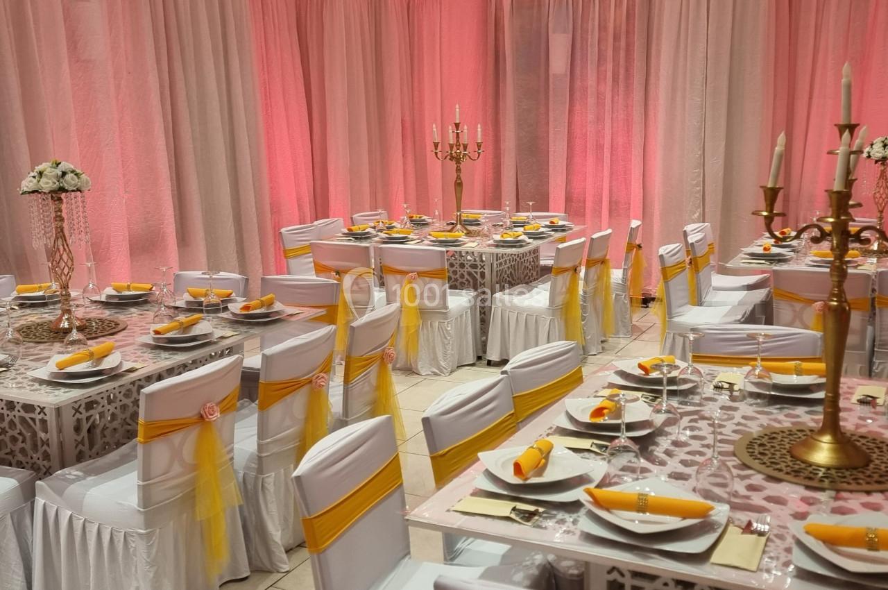 Salle de réception décorée avec des nappes blanches, des chaises ornées de rubans jaunes et des chandeliers dorés.