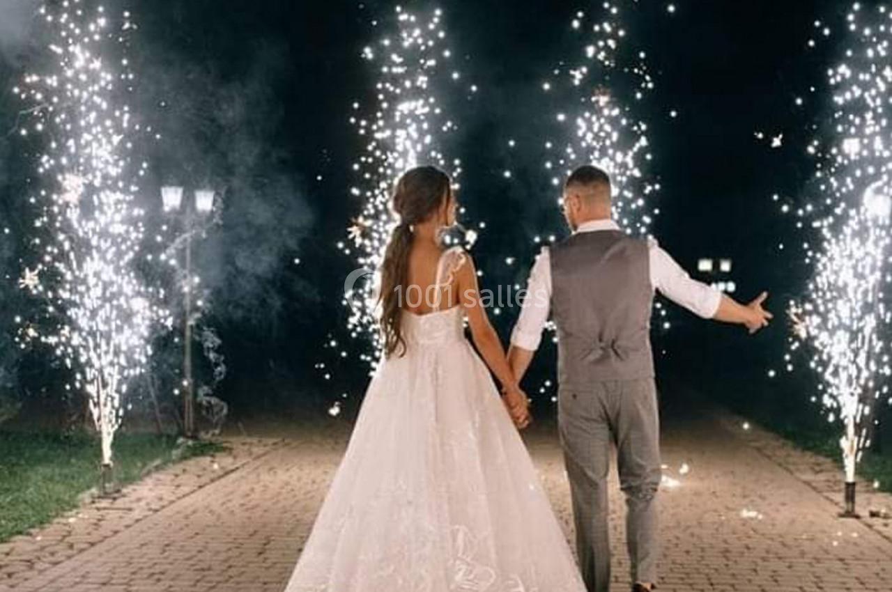Un couple en tenue de mariage marche main dans la main sur une allée illuminée par des feux d'artifice.