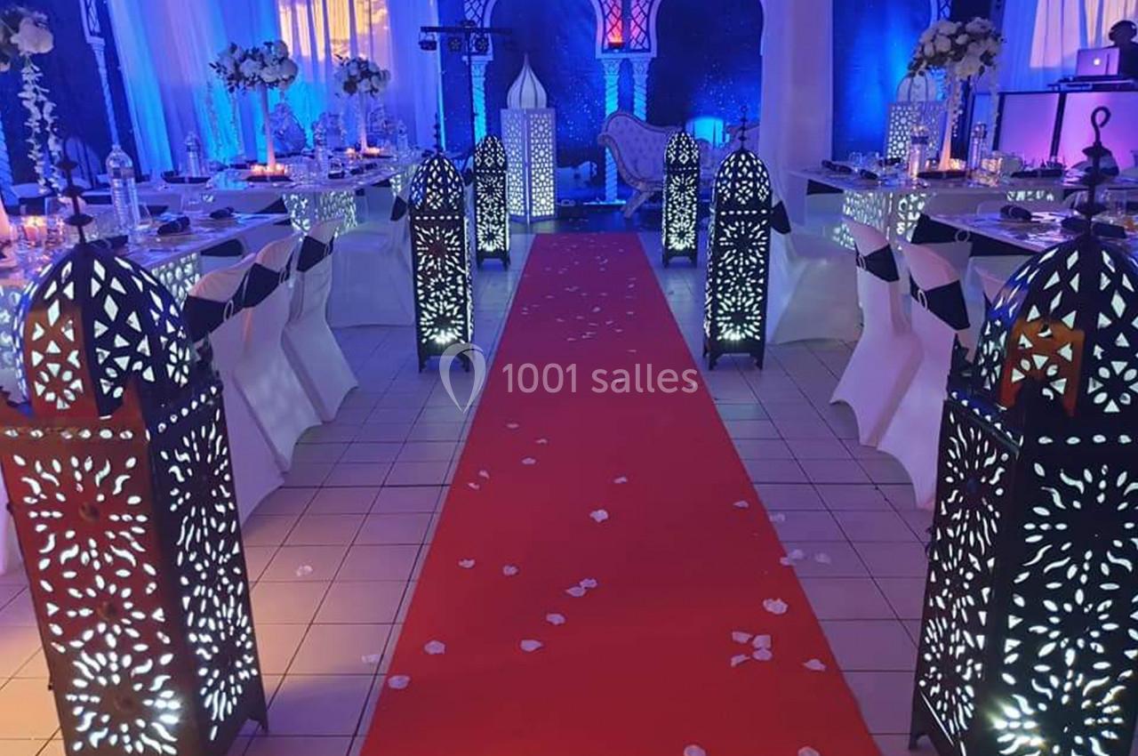 Allée centrale rouge bordée de lanternes décoratives ajourées, entourée de tables dressées dans une salle éclairée.