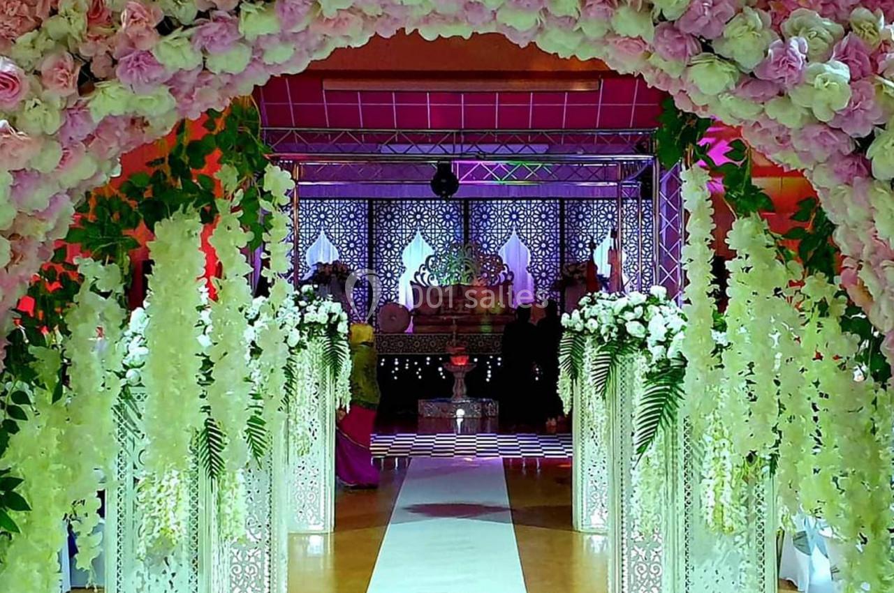 Allée décorée de fleurs blanches et roses menant à une scène ornée de motifs lumineux dans une salle de réception.