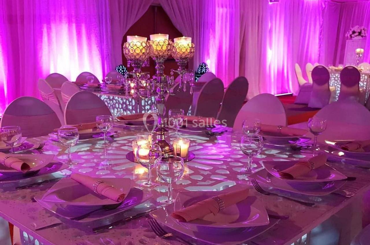 Salle de réception décorée avec des tables élégantes, nappes roses, chandeliers et éclairage tamisé violet.