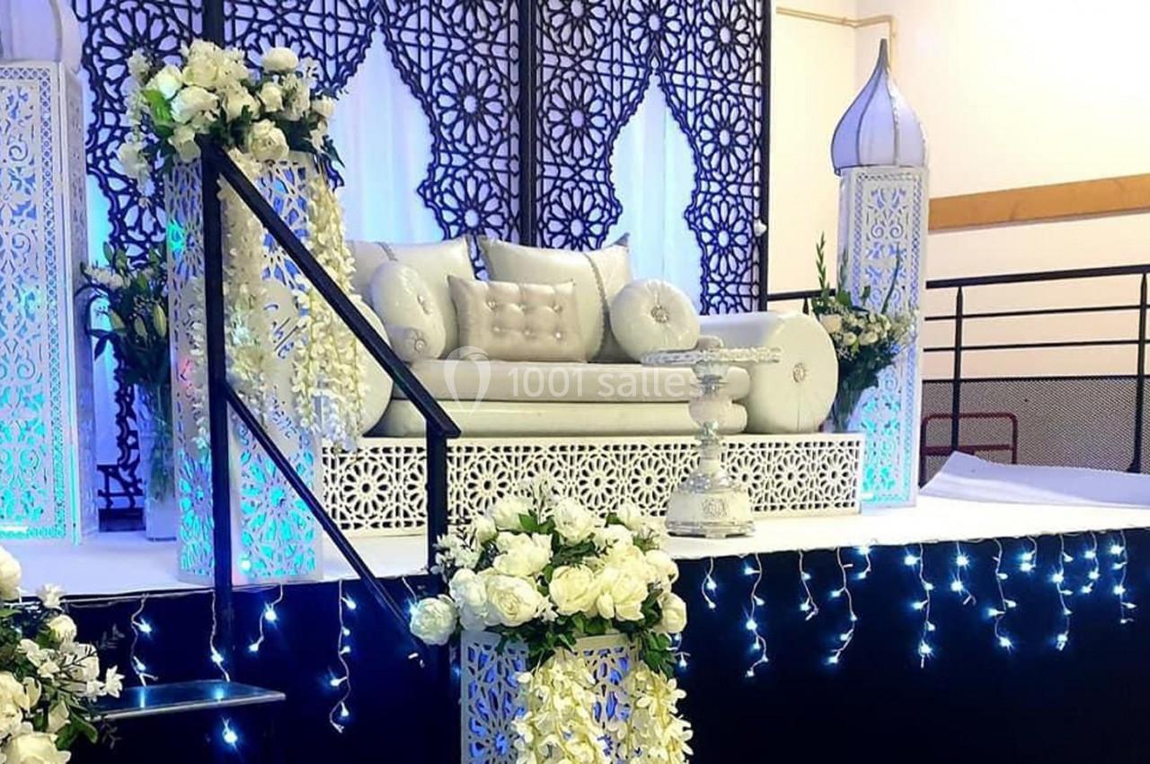 Décor de mariage avec canapé blanc orné de coussins, fleurs blanches et motifs orientaux éclairés par des lumières bleues.