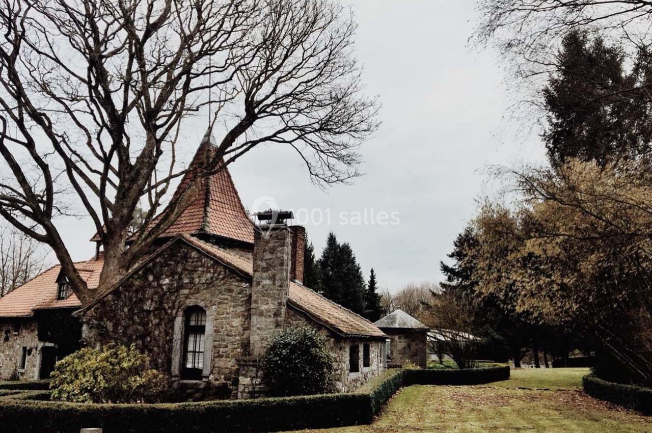 Maison en pierre avec toit en tuiles rouges entourée d'arbres et d'un jardin par temps nuageux.