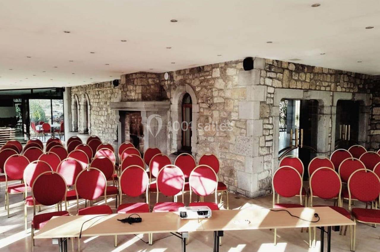 Salle de réunion lumineuse avec murs en pierre, rangées de chaises rouges et tables équipées d'un projecteur.