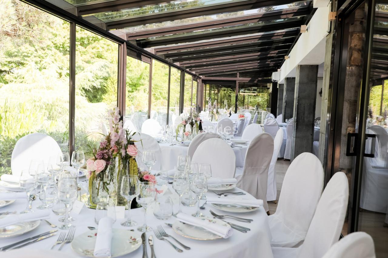 Salle lumineuse avec baies vitrées, tables élégamment dressées avec nappes blanches, fleurs et vaisselle raffinée.