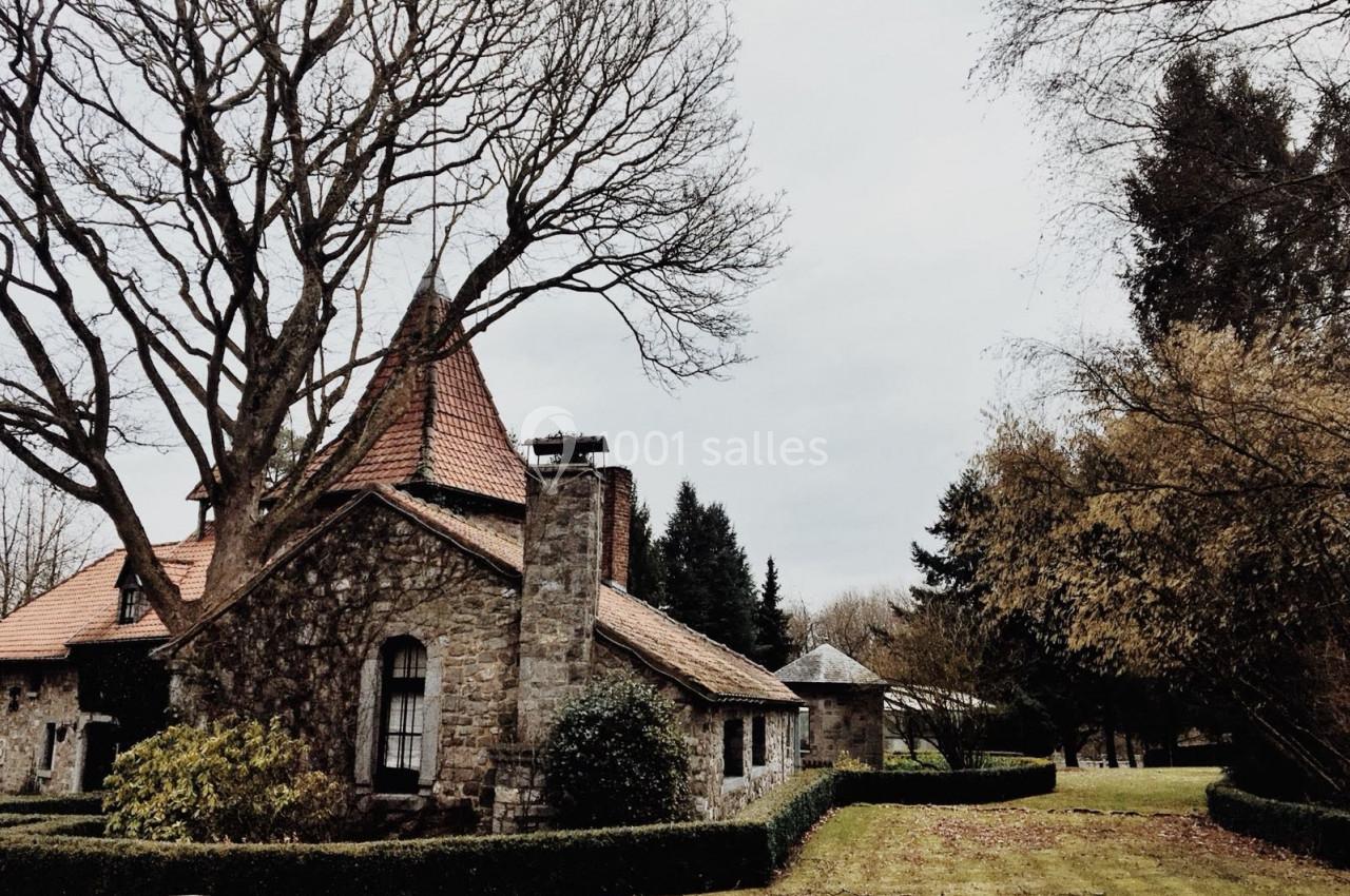 Maison en pierre avec toit en tuiles rouges, entourée d'arbres et d'un jardin parsemé de haies, par temps nuageux.