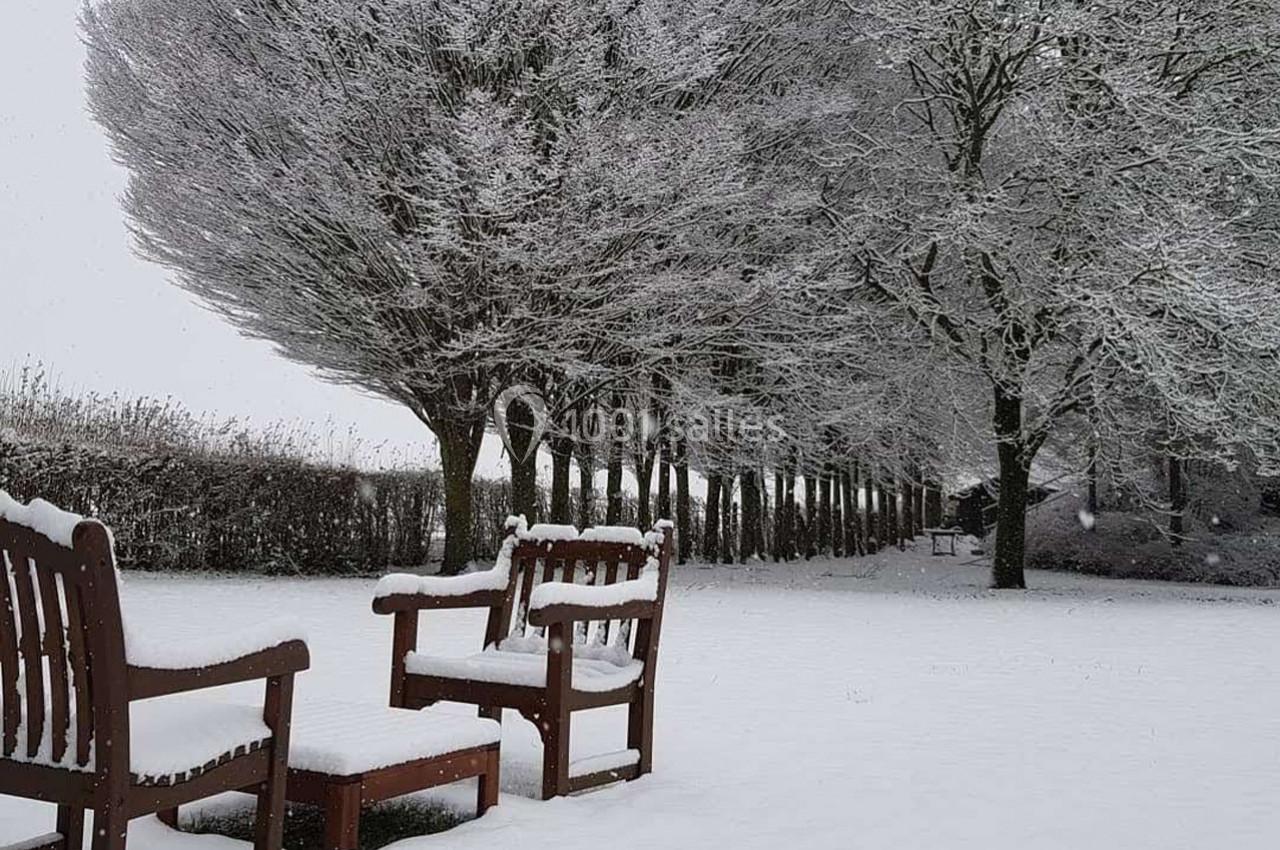 Deux bancs en bois recouverts de neige dans un parc enneigé, entourés d'arbres givrés sous un ciel gris.
