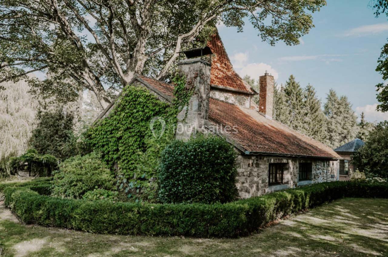 Maison en pierre entourée de verdure, avec un toit en tuiles rouges et des arbres en arrière-plan.