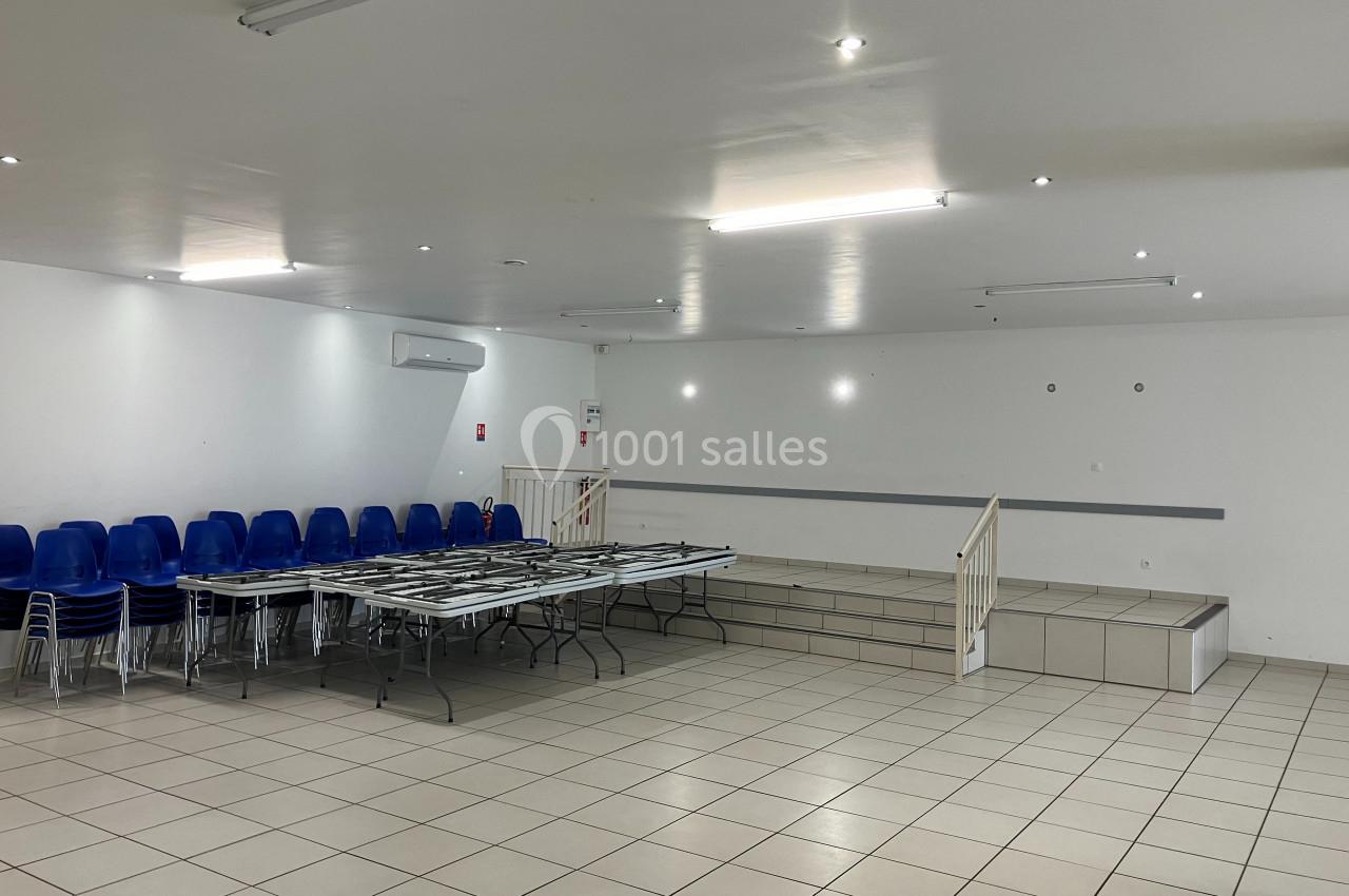 Salle vide avec sol carrelé, tables pliantes, chaises empilées à gauche et petite estrade au fond.