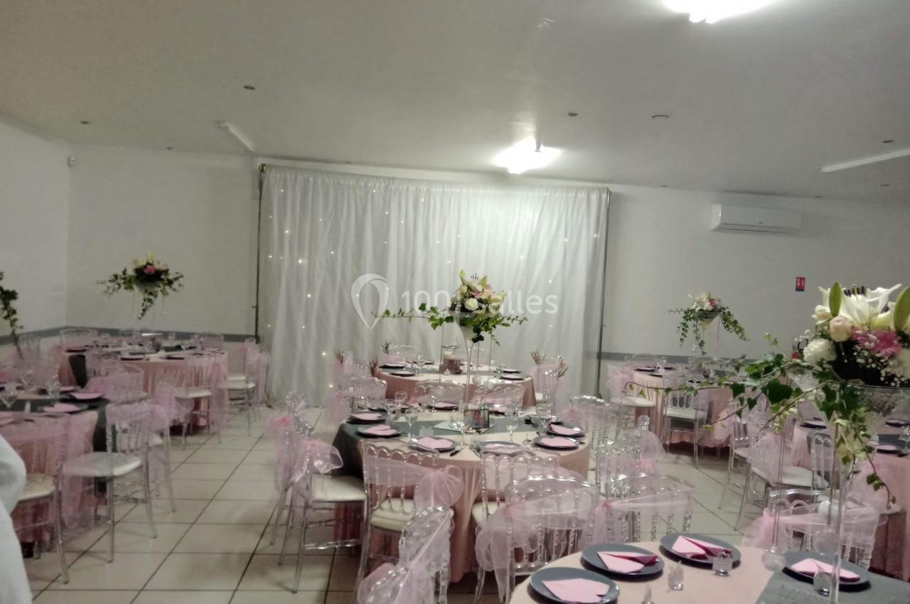 Salle de réception décorée avec des tables rondes, nappes roses, chaises transparentes et arrangements floraux.