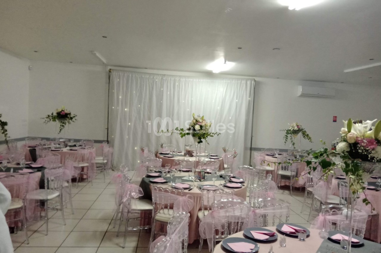 Salle de réception décorée avec des tables rondes, nappes roses, chaises transparentes et arrangements floraux.