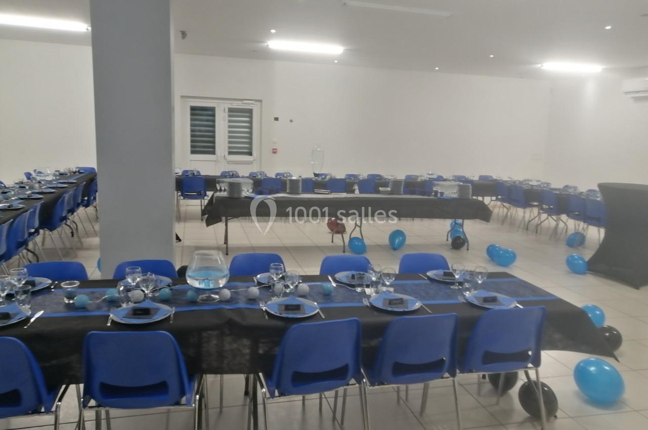 Salle décorée pour un événement avec des tables alignées, nappes noires, vaisselle, et ballons bleus au sol.