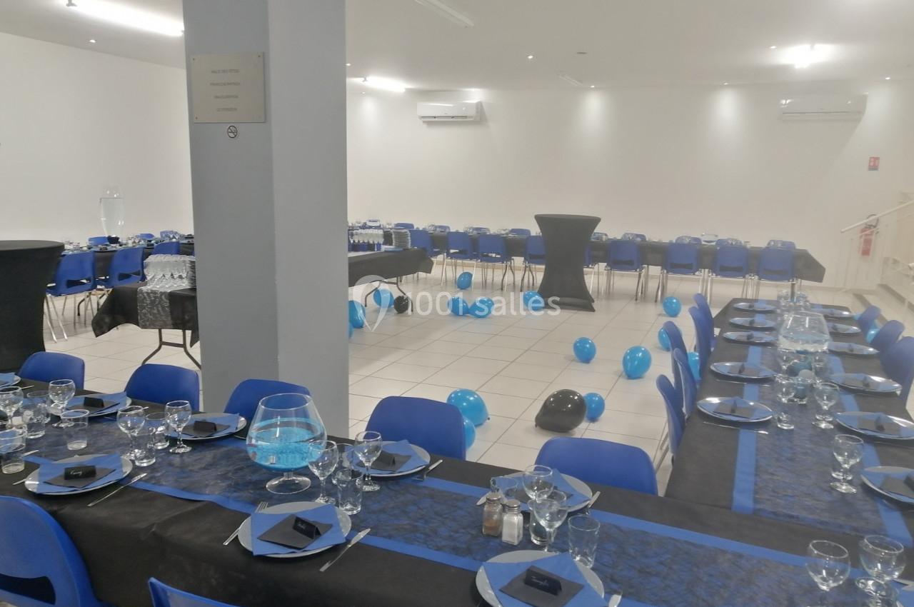 Salle de réception décorée avec des tables dressées en noir et bleu, des ballons au sol et des chaises assorties.
