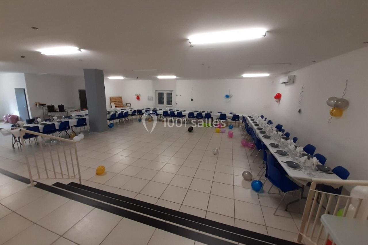 Salle décorée pour un événement, avec tables dressées, chaises bleues et ballons colorés dispersés au sol.