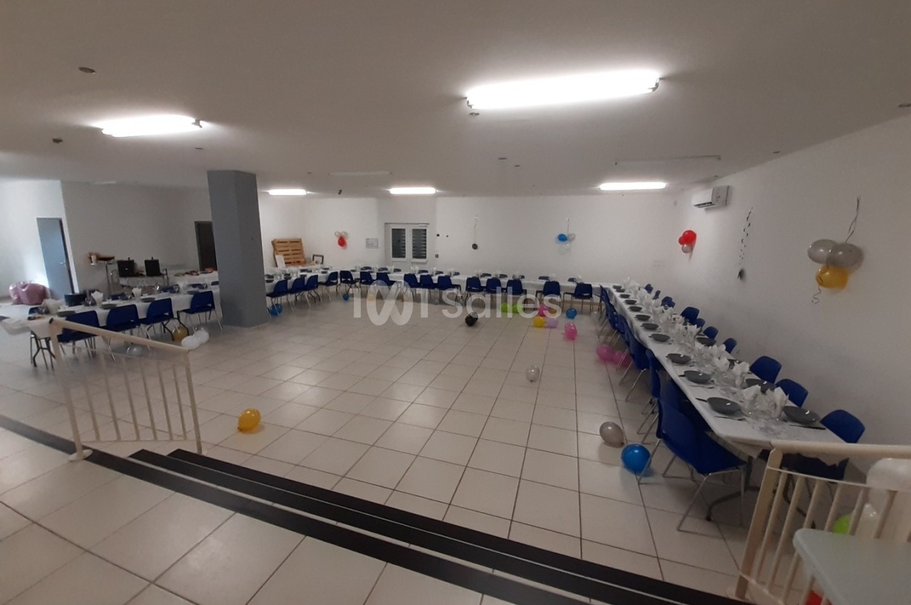 Salle décorée pour un événement, avec tables dressées, chaises bleues et ballons colorés dispersés au sol.