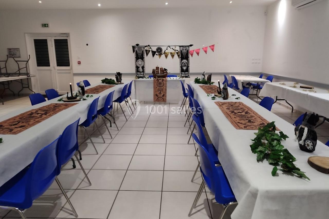 Salle décorée pour un événement, avec tables alignées, nappes blanches, chaises bleues et ornements thématiques.