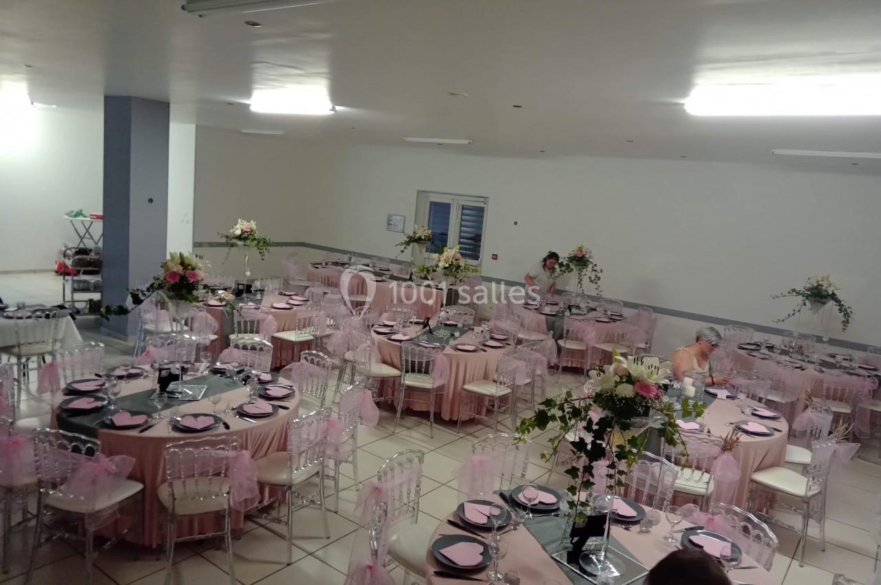 Salle de réception décorée avec des tables rondes, nappes roses, chaises transparentes et arrangements floraux.