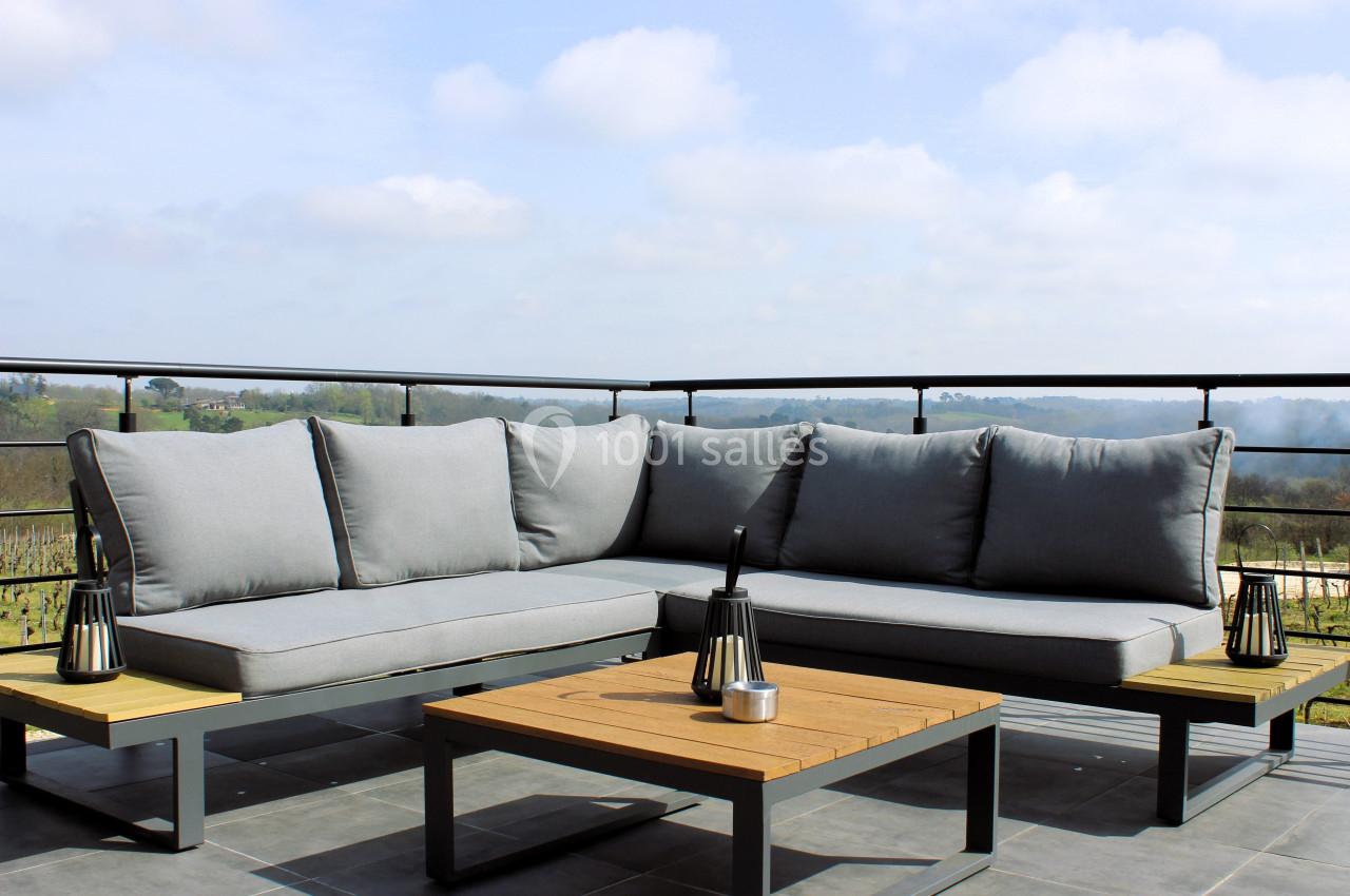 Canapé d'angle gris avec table basse en bois sur une terrasse offrant une vue dégagée sur la campagne.