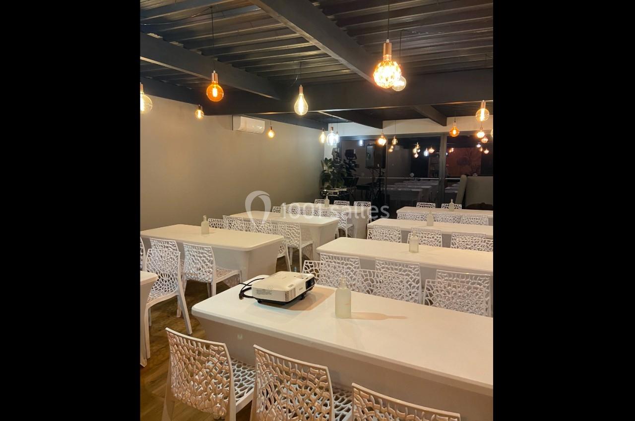 Salle moderne avec tables blanches alignées, chaises ajourées et éclairage suspendu aux ampoules décoratives.