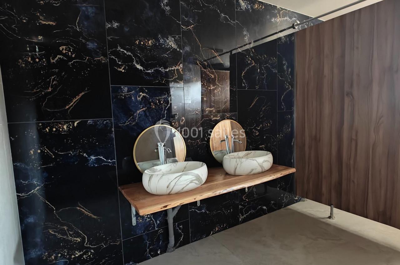 Deux lavabos en marbre avec miroirs ronds sur un plan en bois, devant un mur en carrelage noir à motifs dorés.