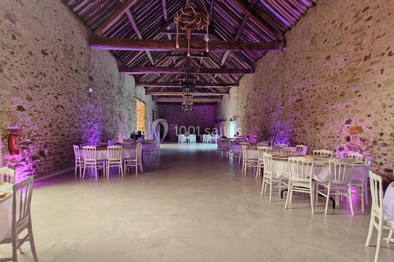 Salle en pierre avec poutres apparentes, tables et chaises blanches disposées pour un événement, éclairage violet.