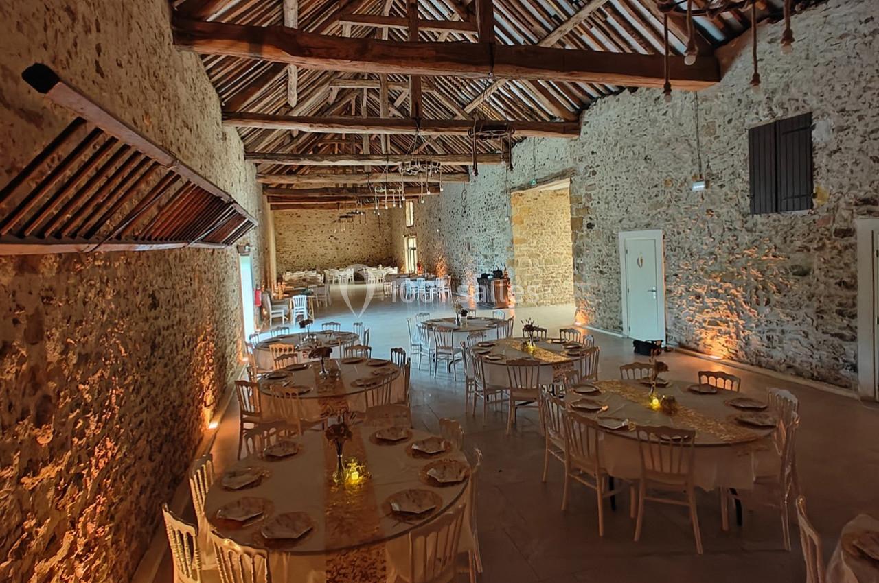 Salle en pierre avec poutres apparentes, tables rondes dressées pour un événement, éclairage chaleureux.