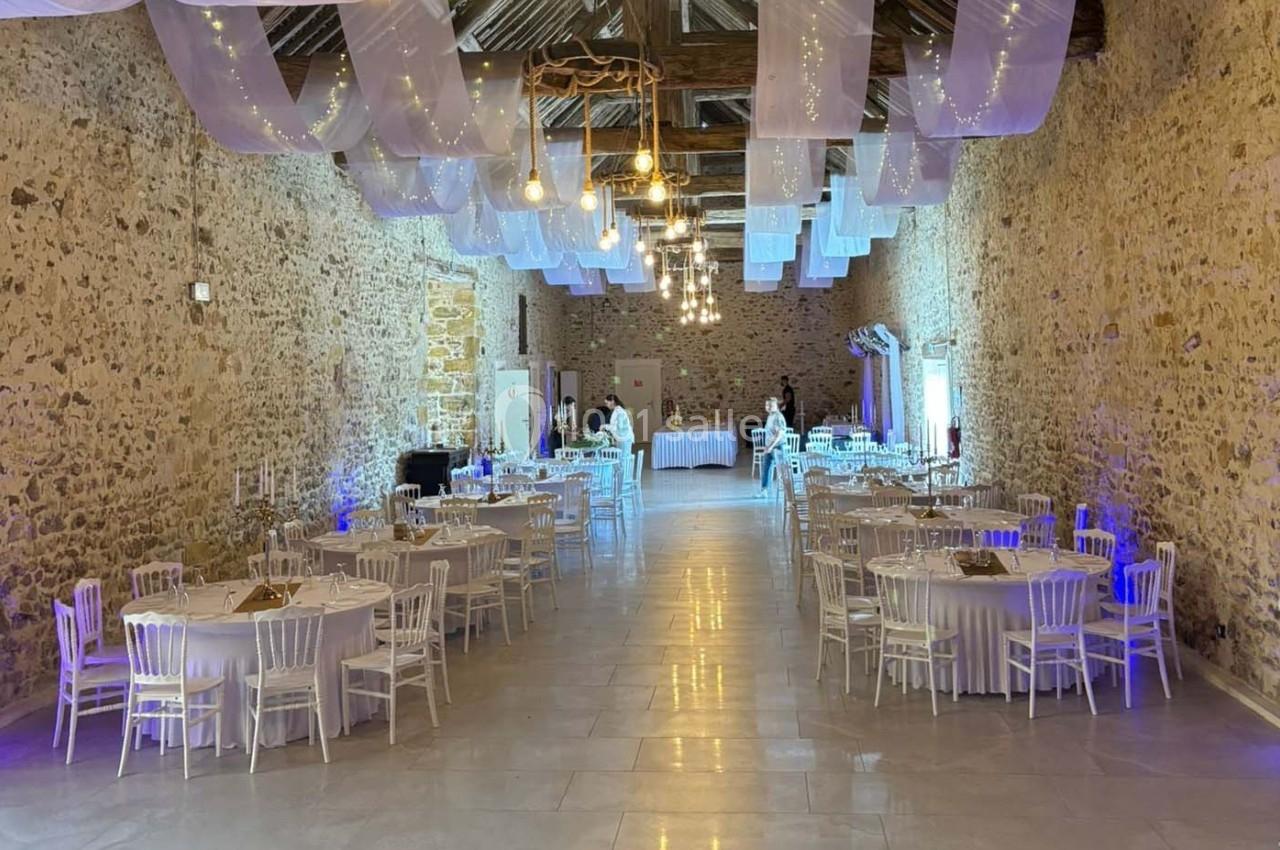 Salle de réception en pierre avec tables rondes dressées, chaises blanches et guirlandes lumineuses suspendues.