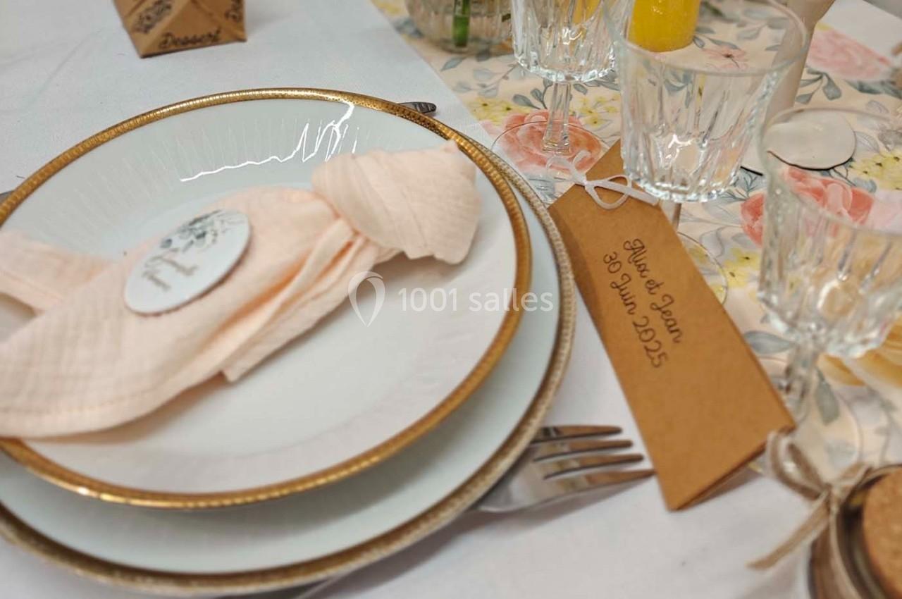 Assiette blanche avec bord doré, serviette rose pliée, et décoration florale sur une table dressée élégamment.