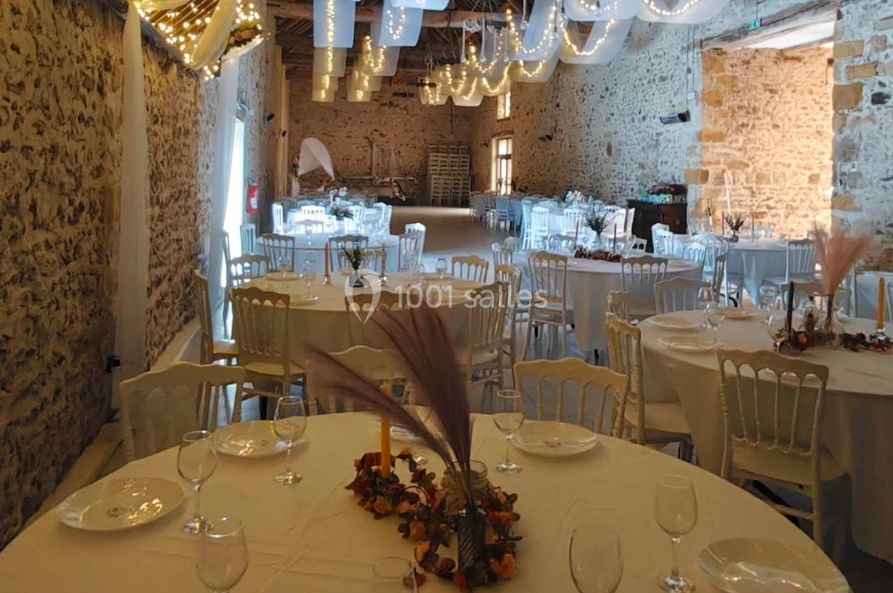 Salle de réception en pierre décorée de guirlandes lumineuses, tables rondes dressées avec nappes blanches et centres de…