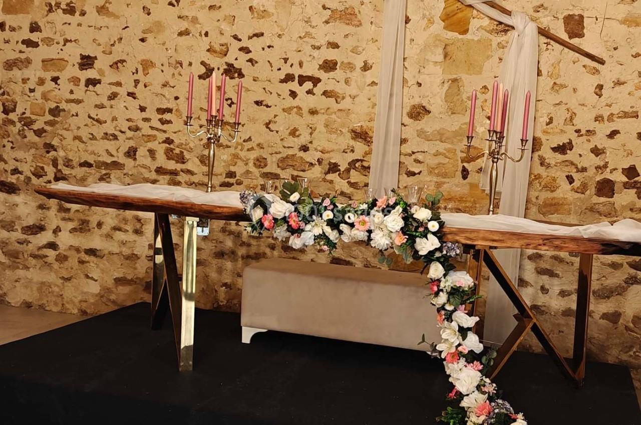 Table en bois décorée de chandeliers roses et de guirlandes florales, placée devant un mur en pierre apparente.