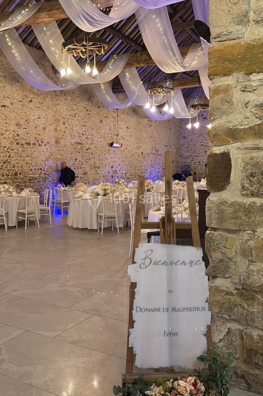 Salle de réception décorée avec des guirlandes lumineuses, des tables dressées et un panneau d'accueil fleuri.