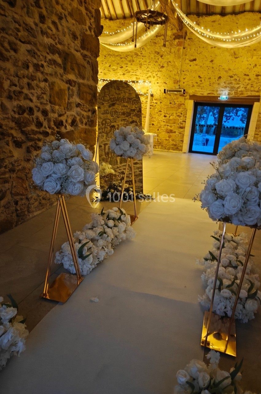 Allée décorée de fleurs blanches dans une salle en pierre éclairée par des guirlandes lumineuses.