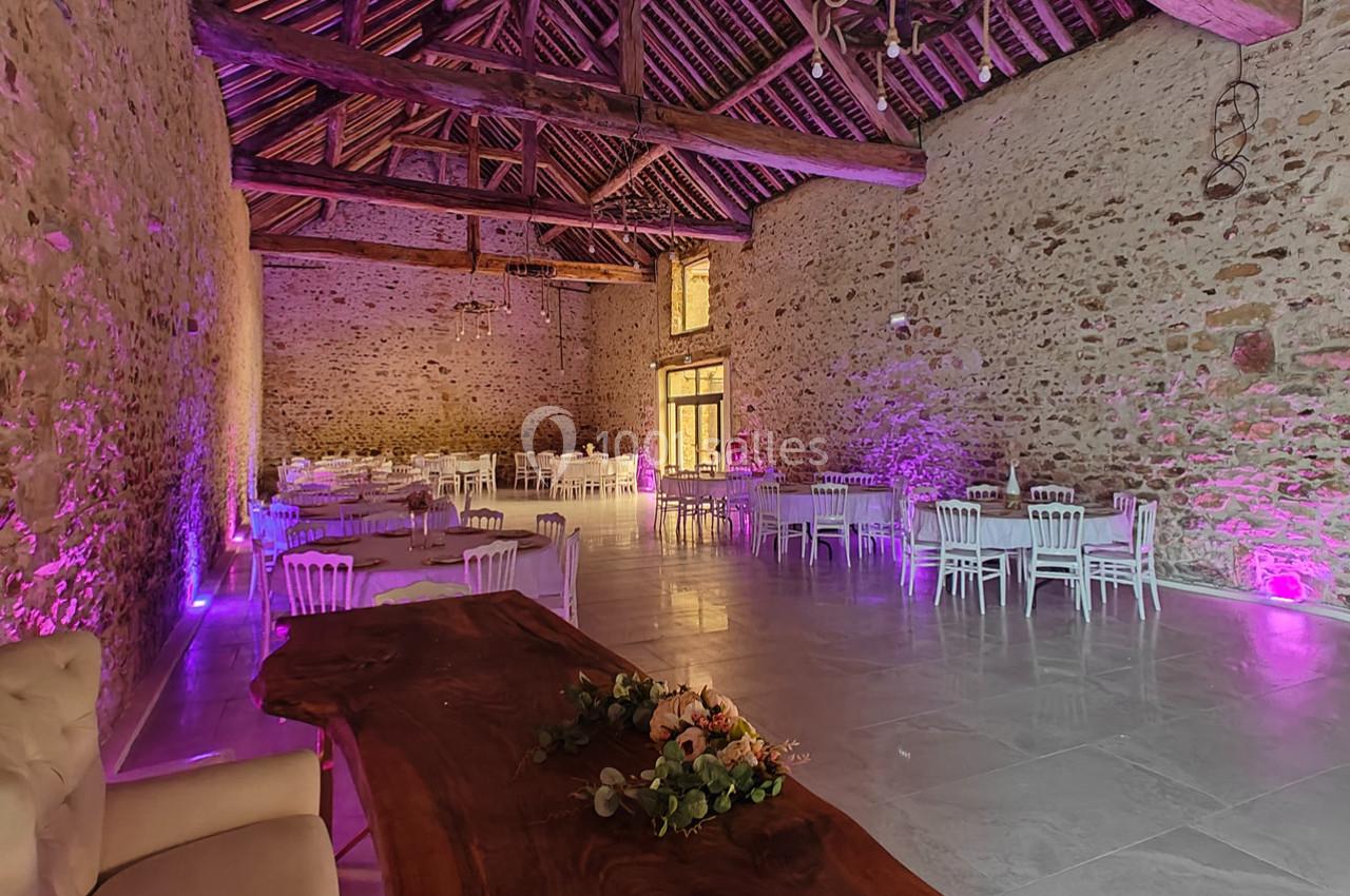 Salle en pierre avec poutres apparentes, tables et chaises blanches, éclairage violet et décoration florale sur une table en…