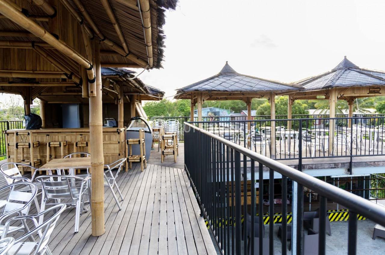 Terrasse en bois avec mobilier en métal, kiosques en bambou et vue sur un parking en arrière-plan.