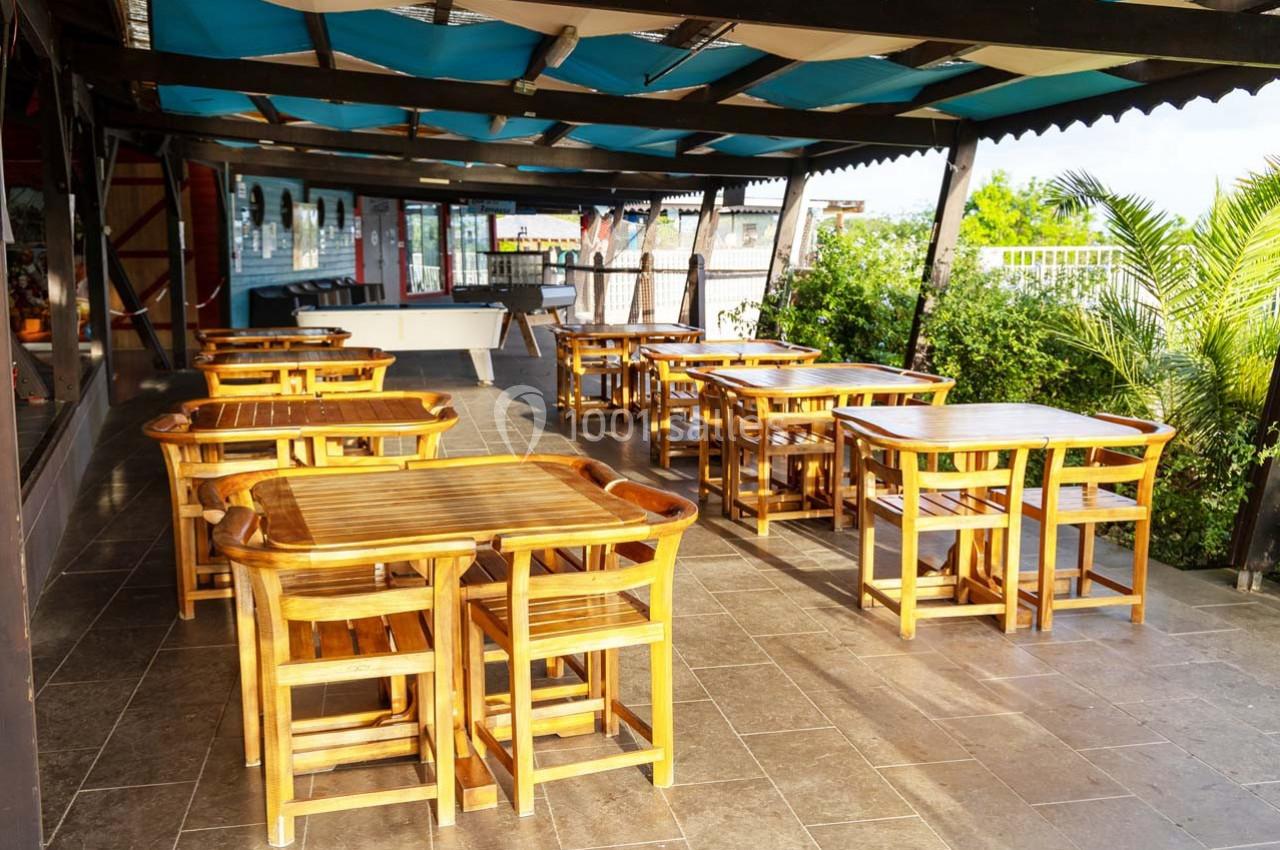 Terrasse couverte avec des tables et chaises en bois, entourée de verdure et éclairée par la lumière naturelle.