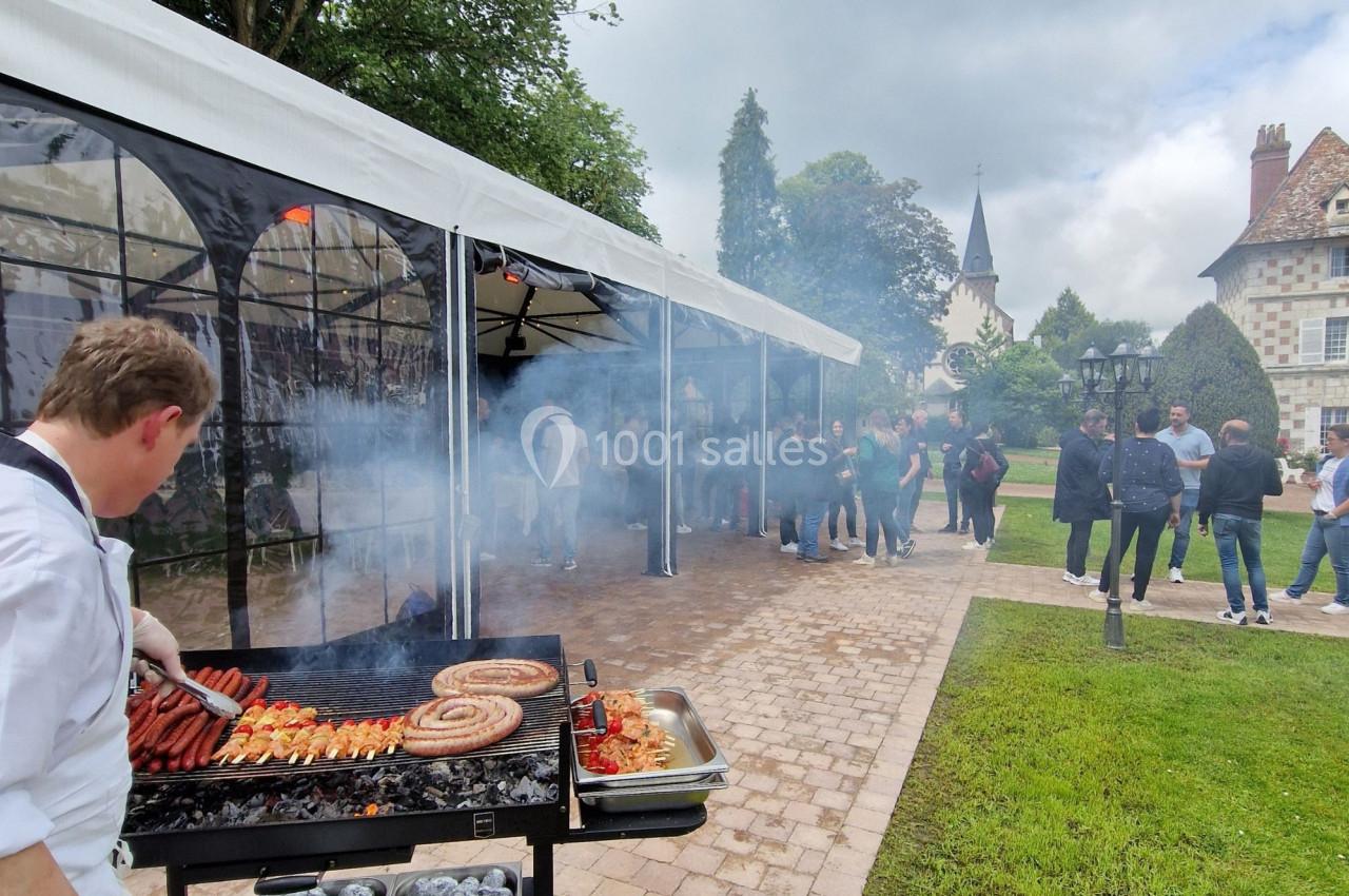 Un cuisinier prépare des grillades sur un barbecue lors d'un événement en plein air, avec des invités en arrière-plan.
