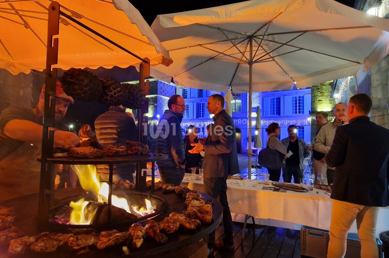 Des convives discutent près d'un barbecue en plein air, sous des parasols éclairés, dans une ambiance nocturne festive.