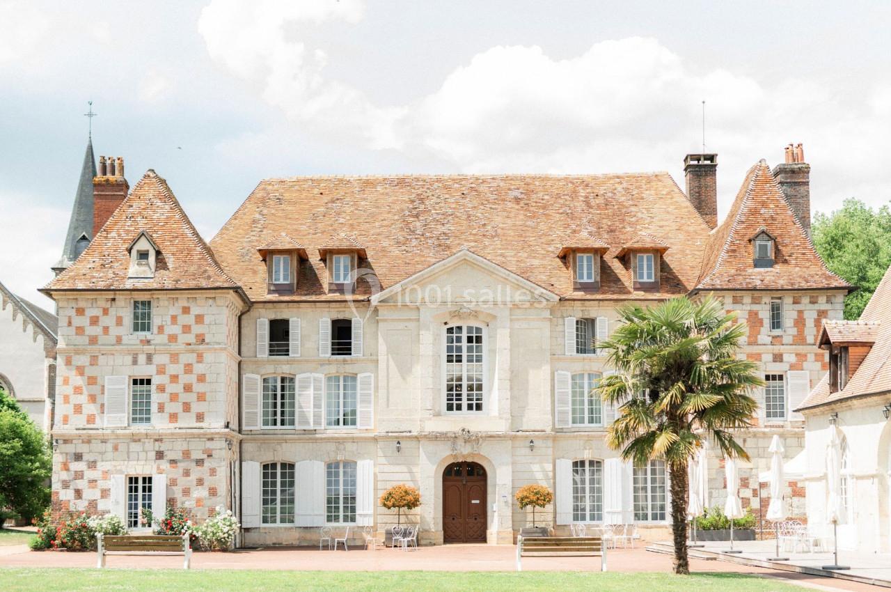Façade d'un manoir en pierre et briques avec toit en tuiles, entouré de verdure et d'une cour dégagée.