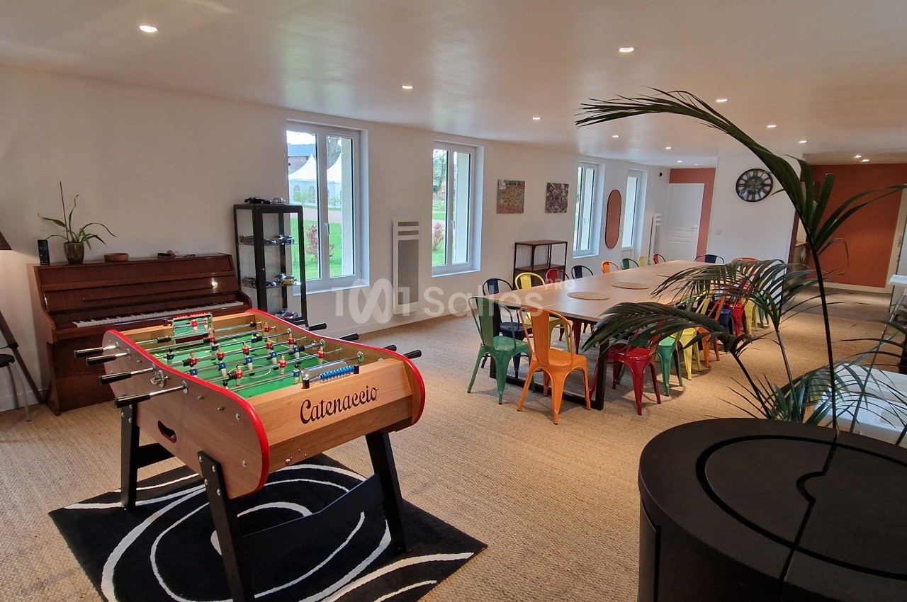 Salle lumineuse avec baby-foot, grande table entourée de chaises colorées, et vue sur un jardin à travers les fenêtres.