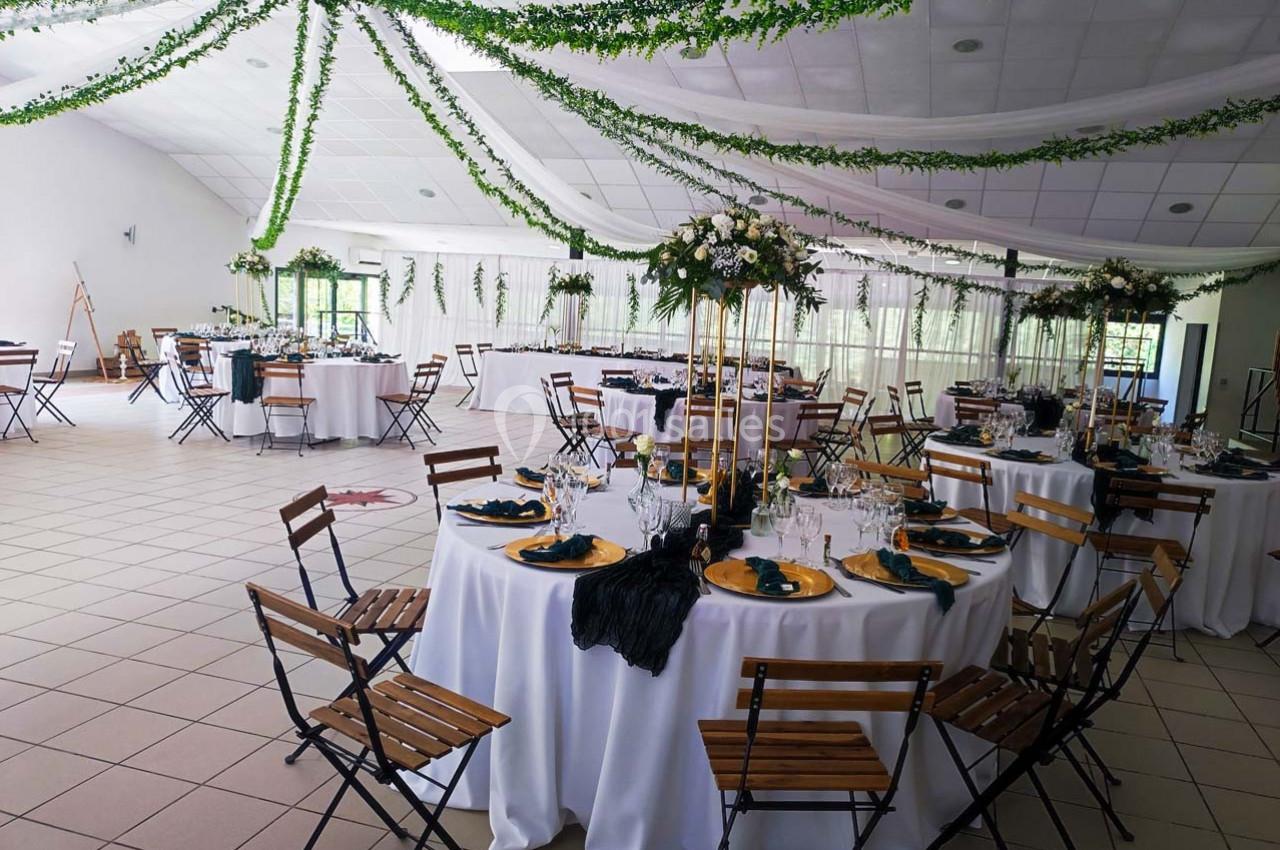 Salle de réception décorée avec des tables rondes, nappes blanches, chaises en bois et guirlandes végétales suspendues.