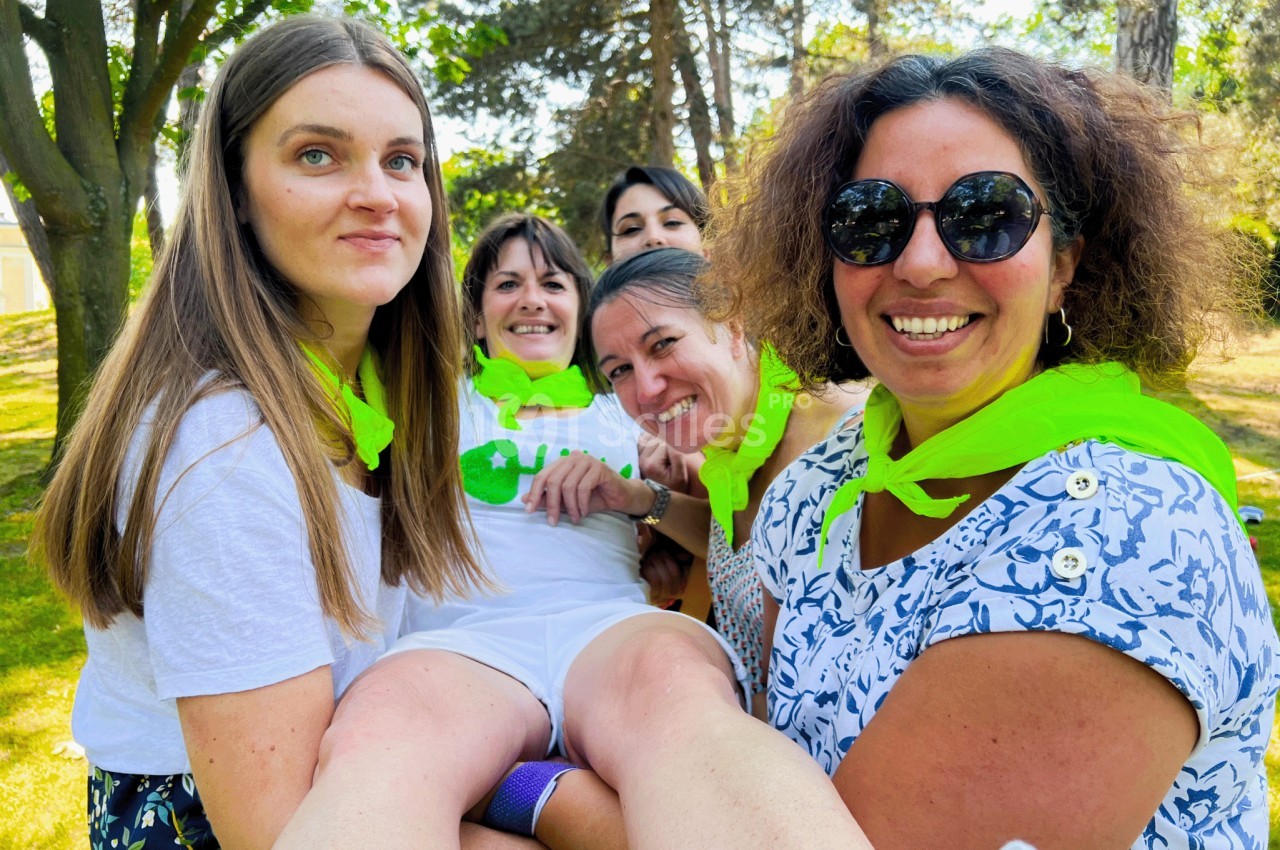 Cinq femmes souriantes portant des foulards verts, réunies dans un parc ensoleillé.