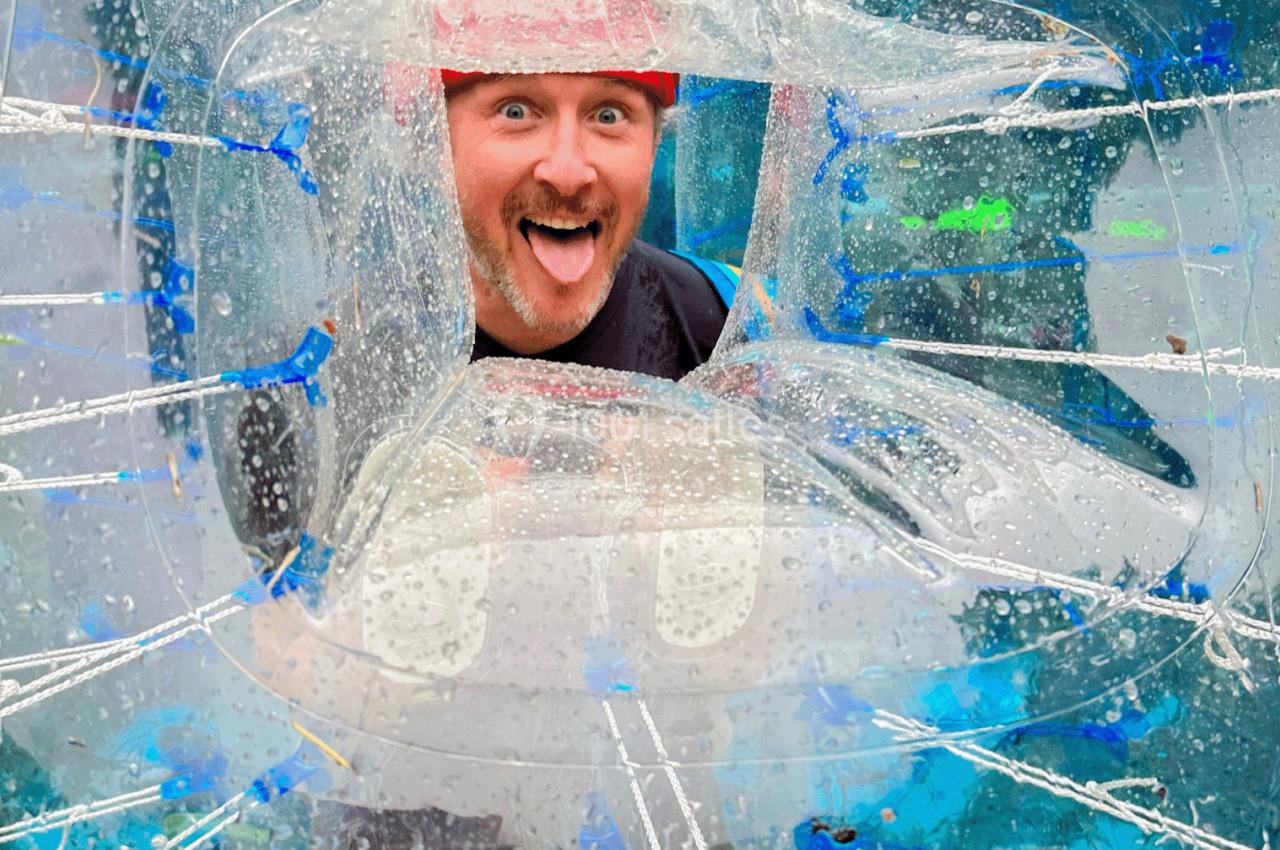 Homme souriant à travers une bulle gonflable transparente, portant un casque rouge, dans un cadre extérieur.