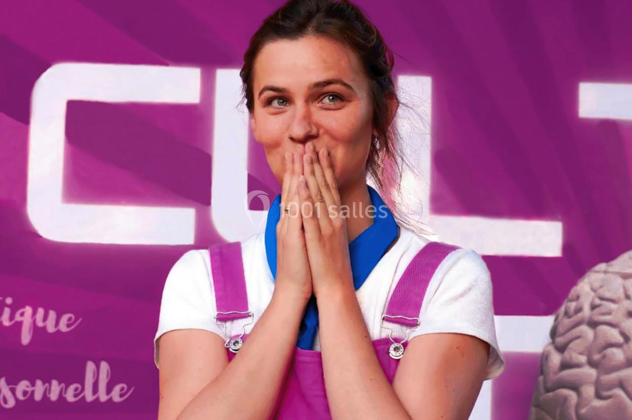 Femme souriante en salopette rose, mains sur le visage, devant un décor violet avec un cerveau en arrière-plan.