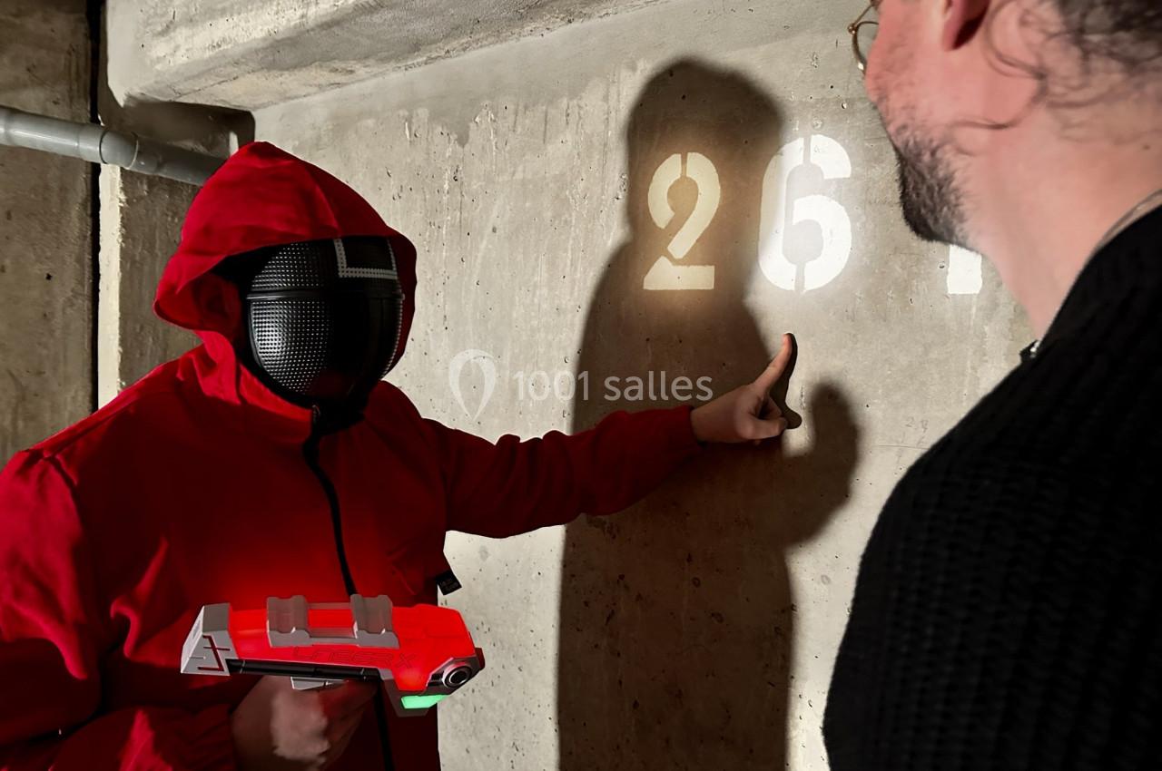 Une personne en rouge avec un masque pointe un mur marqué ’267’ dans un environnement sombre, tenant un pistolet jouet.