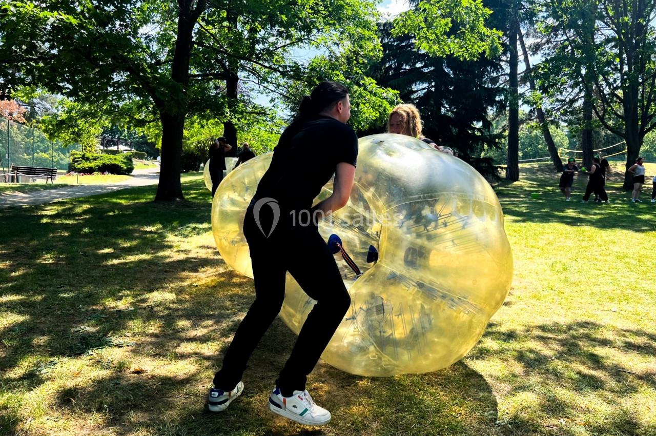 Deux personnes jouent au bubble football dans un parc ensoleillé entouré d'arbres.